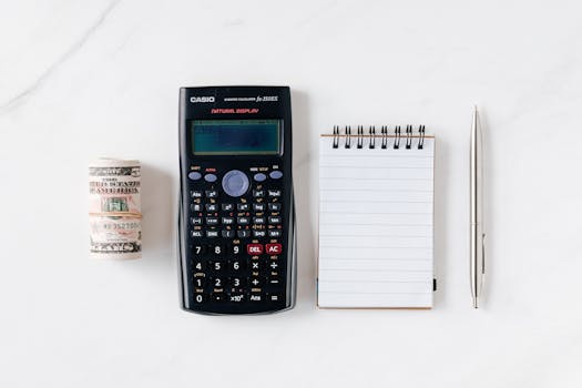 Top view of financial tools including a calculator, notepad, pen, and rolled cash on a white background.